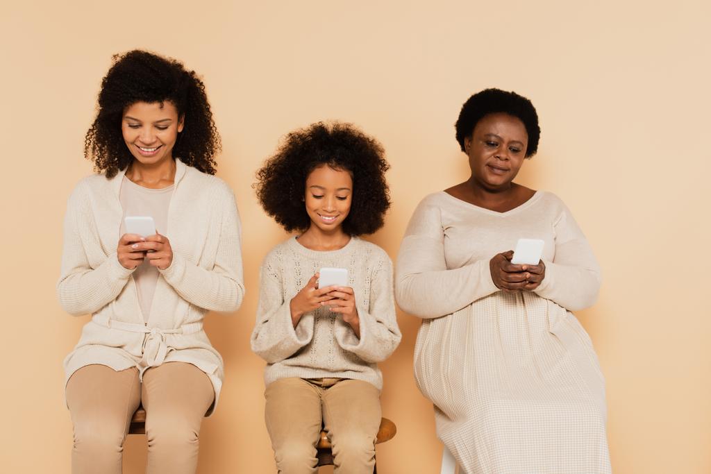 Αφροαμερικανή κόρη, εγγονή και γιαγιά κάθονται και κοιτάζουν τα κινητά τηλέφωνα σε μπεζ φόντο - Φωτογραφία, εικόνα