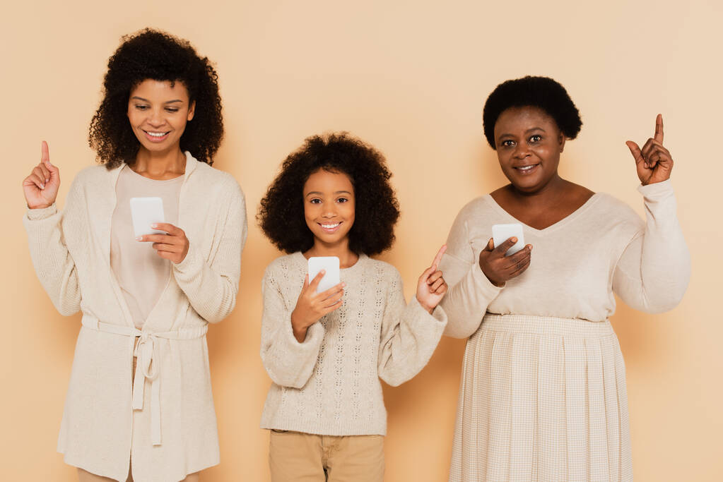 afro-amerikai lánya, unokája és nagymamája ujjal felfelé mutató és kezében mobiltelefonok bézs alapon - Fotó, kép