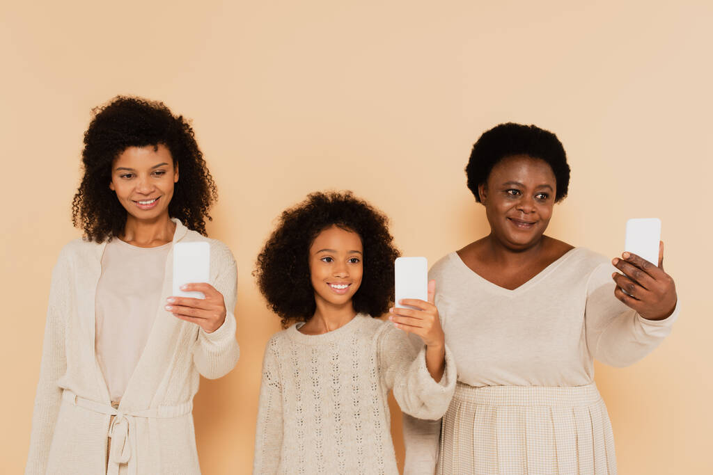 Αφροαμερικανή κόρη, εγγονή και γιαγιά βγάζουν selfies στα κινητά σε μπεζ φόντο - Φωτογραφία, εικόνα