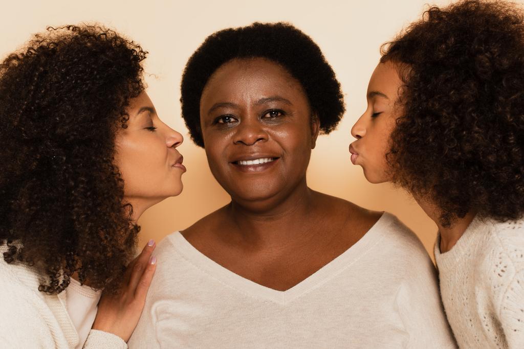 afrikkalainen amerikkalainen tytär ja tyttärentytär suutelevat isoäitiä suljetuin silmin beige taustalla - Valokuva, kuva