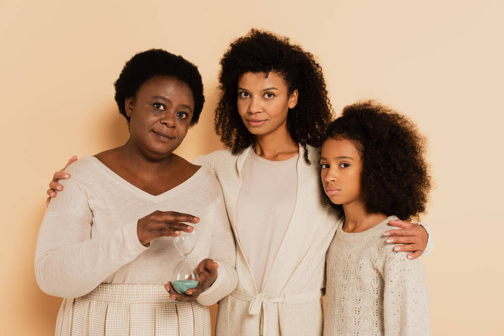 serious african american grandmother holding sandglass with daughter and granddaughter standing nearby on beige background - Photo, Image