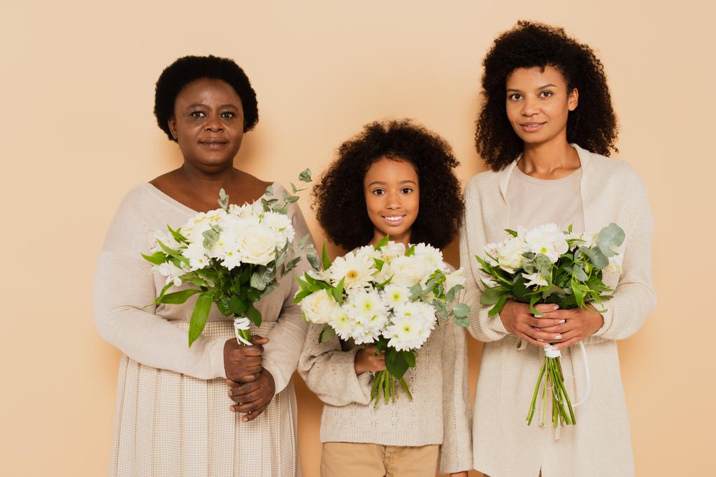 ευτυχισμένη οικογένεια αφροαμερικανών γιαγιά, κόρη και εγγονή με ανθοδέσμες των λουλουδιών που απομονώνονται σε μπεζ - Φωτογραφία, εικόνα