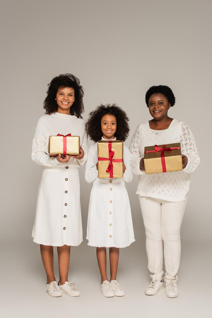 πλήρους μήκους πλάνο της χαμογελαστή Αφροαμερικανή μητέρα, κόρη και γιαγιά κρατώντας κουτιά δώρων σε γκρι φόντο - Φωτογραφία, εικόνα