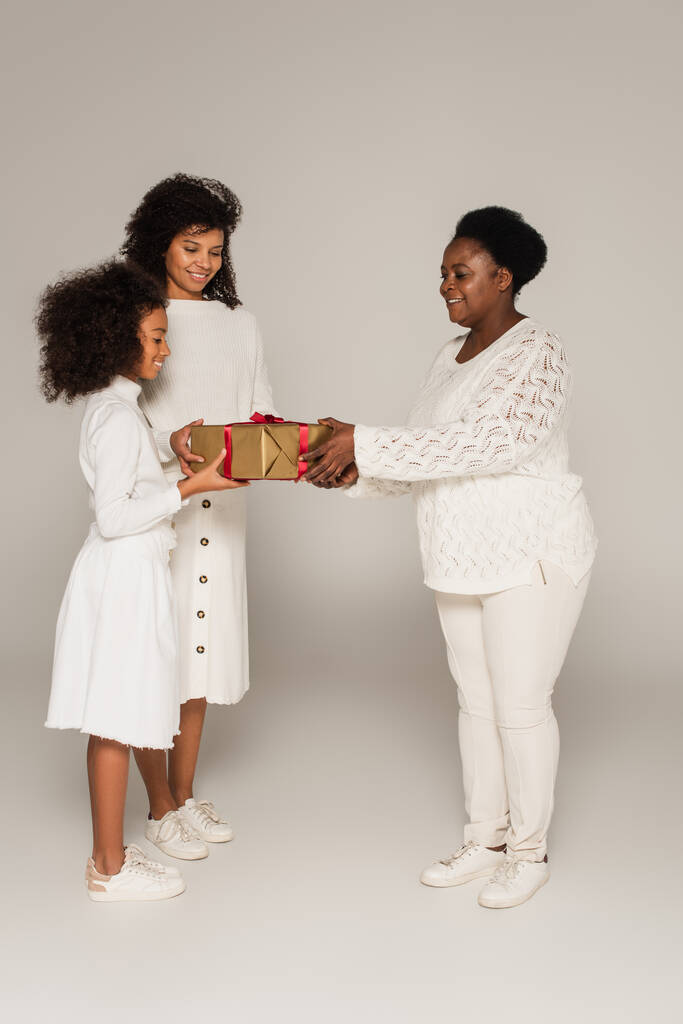 teljes hosszúságú lövés mosolygós afro-amerikai nagymama bemutató ajándék doboz lánya és unokája szürke háttér - Fotó, kép