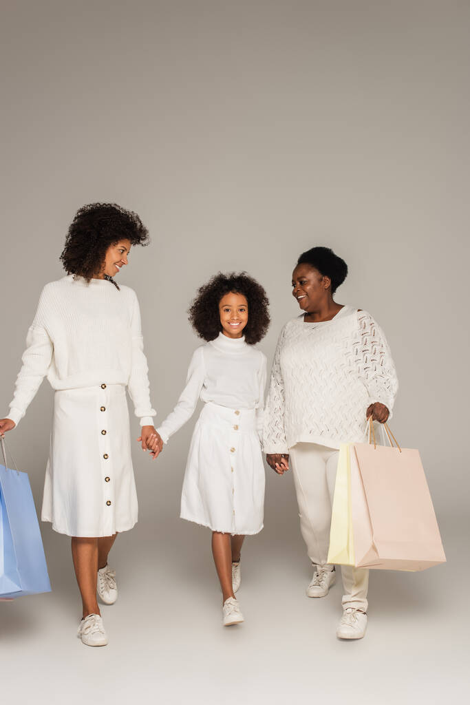 mosolygós afro-amerikai anya, lánya és nagymamája séta bevásárló táskák és kézen fogva szürke háttér - Fotó, kép