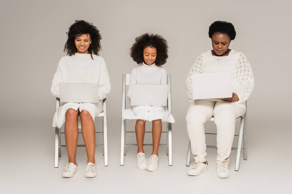 Αφροαμερικανή κόρη, μητέρα και γιαγιά κάθονται σε καρέκλες και κοιτάζουν φορητούς υπολογιστές σε γκρι φόντο - Φωτογραφία, εικόνα