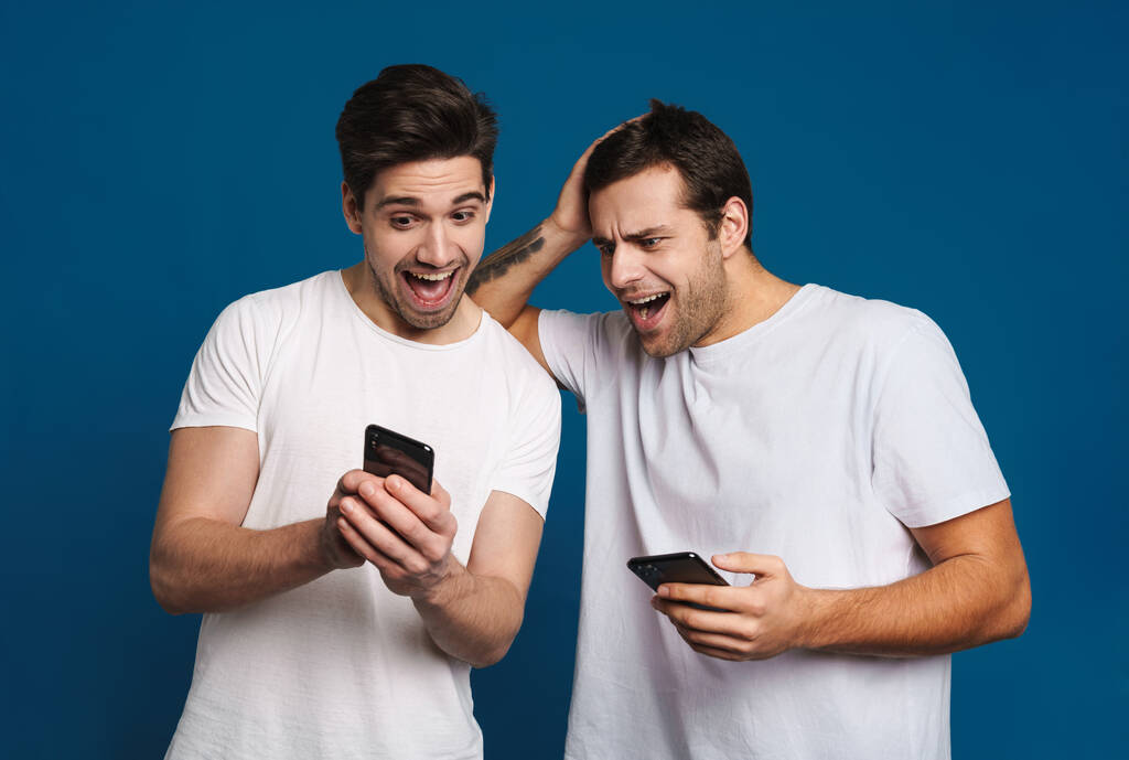 Encantado de dos chicos que expresan sorpresa mientras posan con teléfonos inteligentes aislados sobre fondo azul - Foto, imagen