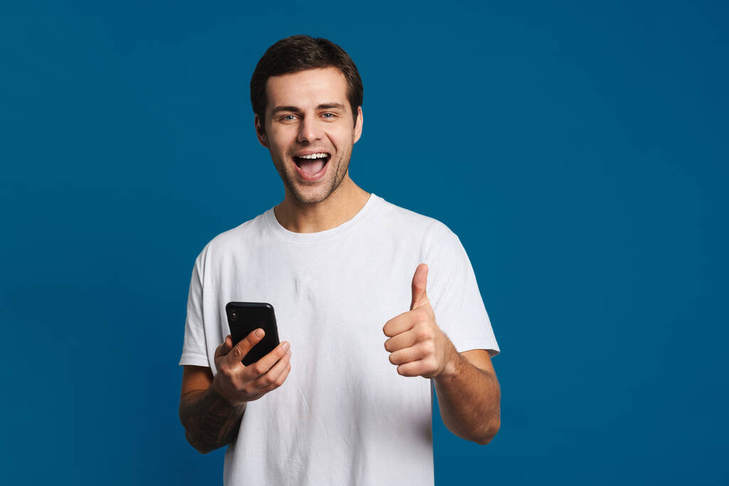 Щасливий незахищений хлопець показує великий палець під час використання мобільного телефону ізольовано на синьому фоні
 - Фото, зображення