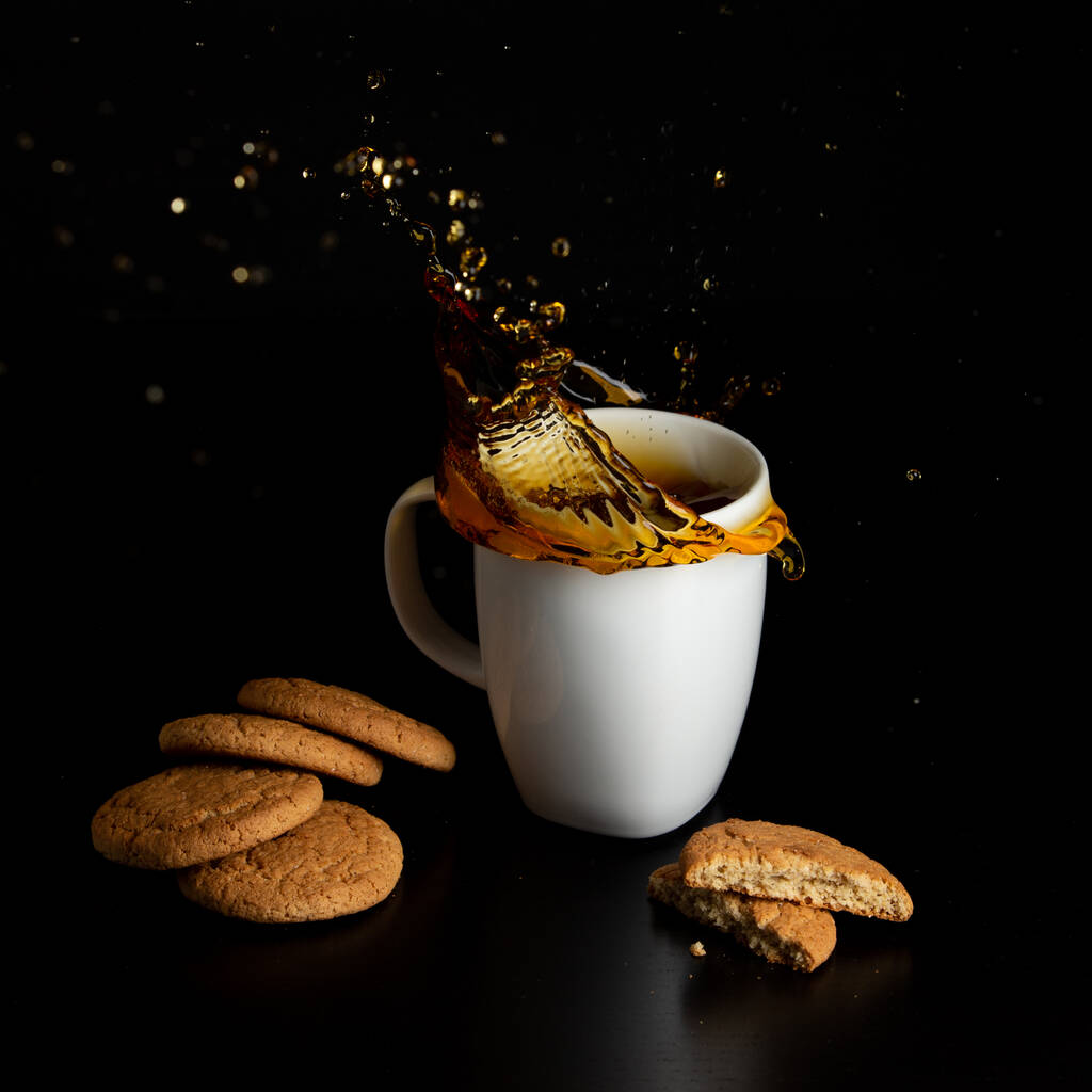 Glass with tea pouring with liquid with splashes and drops of water. Brewing tea concept. Cup pouring with water or tea with splashes on dark wooden background. Cup, teabag and cookies on table. - Photo, Image