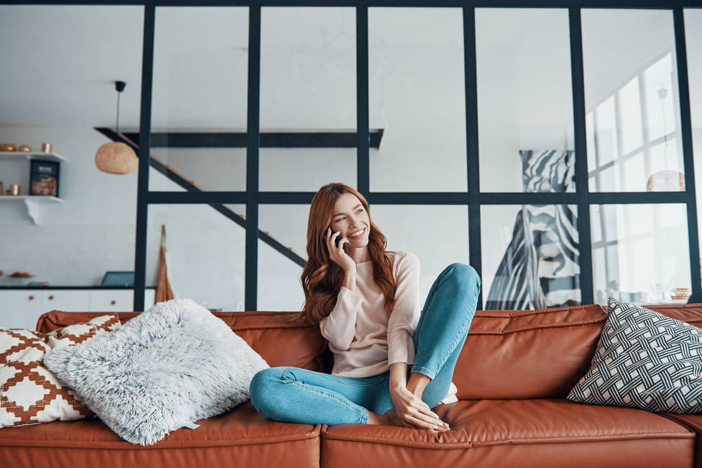 Улыбающаяся молодая женщина берет трубку, сидя дома на диване - Фото, изображение