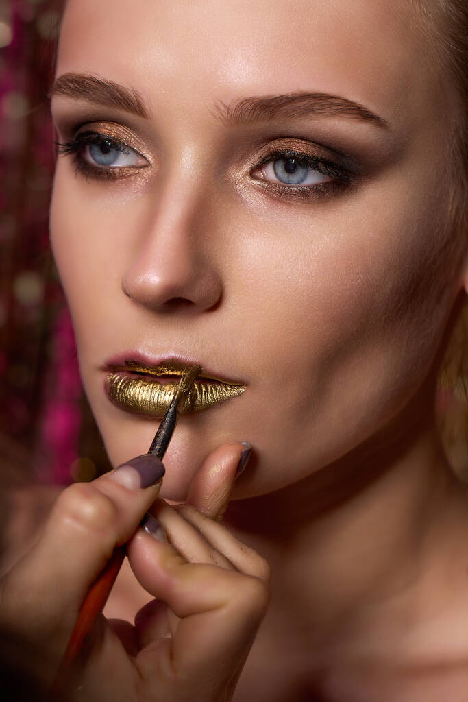 Joven hermosa chica aplicando maquillaje labios de oro por maquillaje artista de cerca - Foto, Imagen