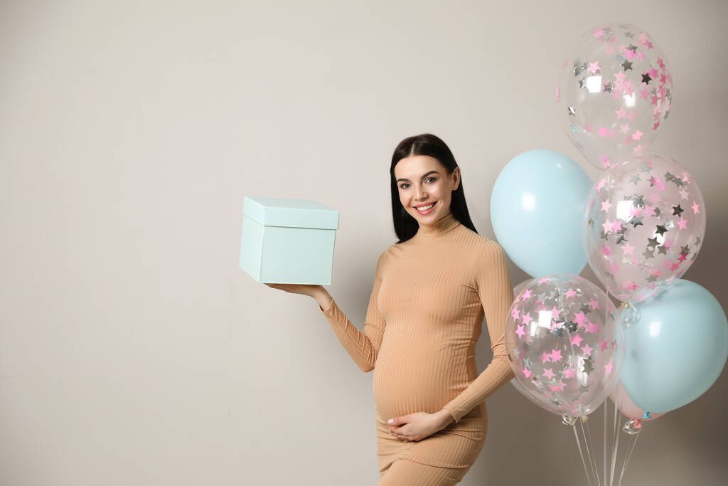 Mujer embarazada feliz con caja de regalo y globos cerca de la pared gris, espacio para el texto. Fiesta de ducha bebé - Foto, imagen
