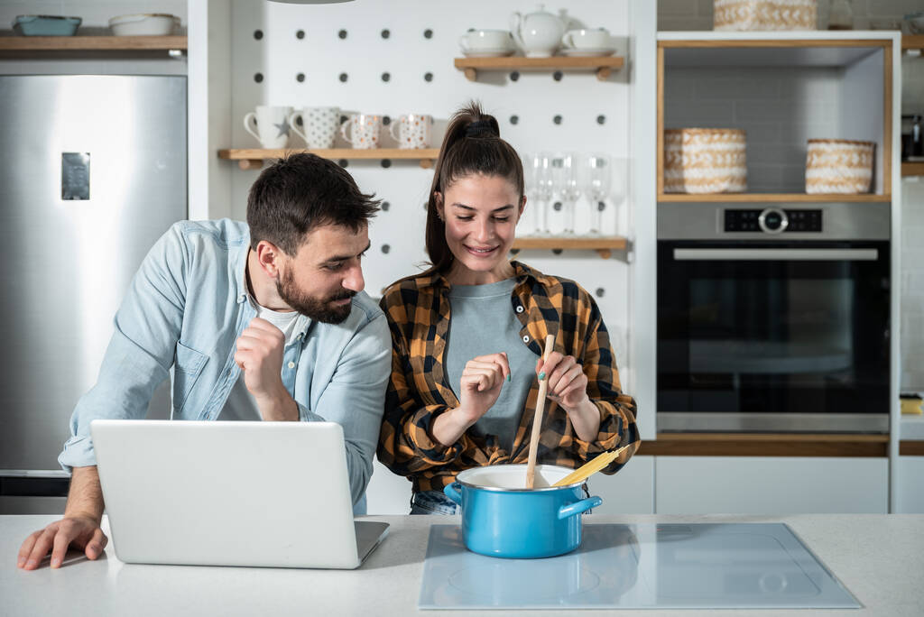 Joven feliz pareja en la cocina hombre burlas de su novia cómo ella no sabe cocinar cuando ella necesita ordenador portátil y utilizar recetas en línea de Internet - Foto, Imagen
