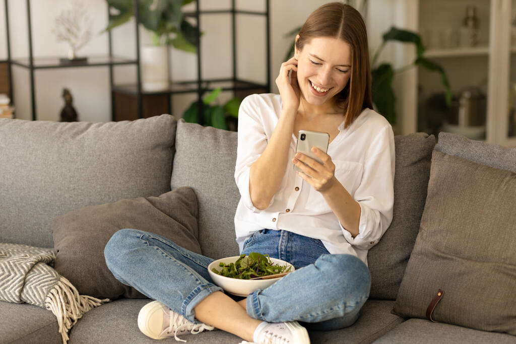 Uśmiechnięta młoda, kaukaska milenijna kobieta rozmawiająca w sieciach społecznościowych za pomocą aplikacji mobilnych, siedząca na kanapie w domu - Zdjęcie, obraz