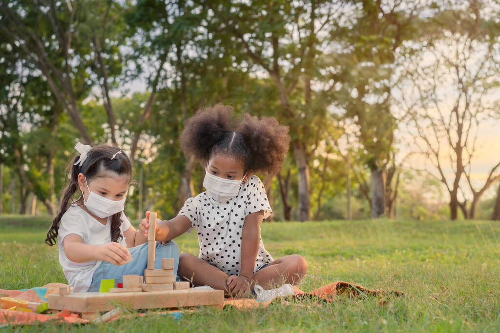 Fröhlich fröhliche ethnische Mädchen spielen gemeinsam Holzklötze Puzzle im Freien Park, Sie tragen Masken, um sie vor Viren zu schützen, Beziehung kleine Kinder, Diverse ethnische Konzept. - Foto, Bild