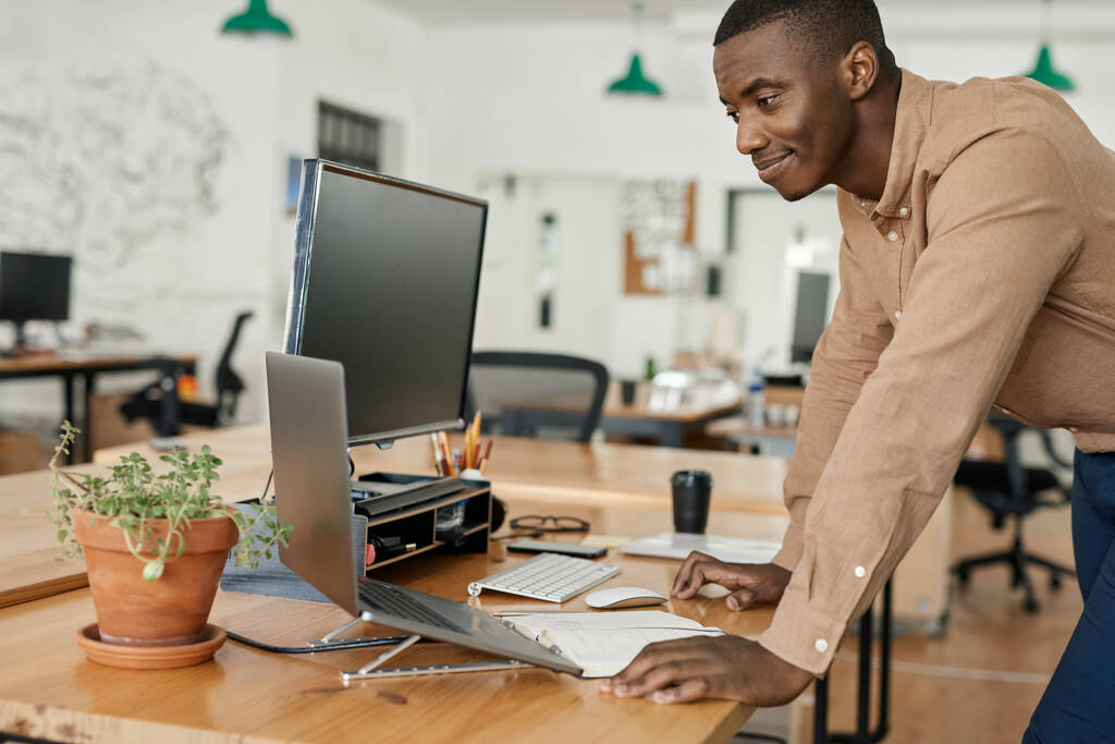 Χαμογελώντας νεαρός Αφρικανός επιχειρηματίας που γέρνει πάνω από το γραφείο του σε ένα σύγχρονο γραφείο και εργάζεται σε ένα φορητό υπολογιστή - Φωτογραφία, εικόνα
