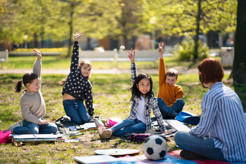 Nauczyciel z małymi dziećmi siedzącymi na świeżym powietrzu w parku miejskim, koncepcja kształcenia grupowego. - Zdjęcie, obraz