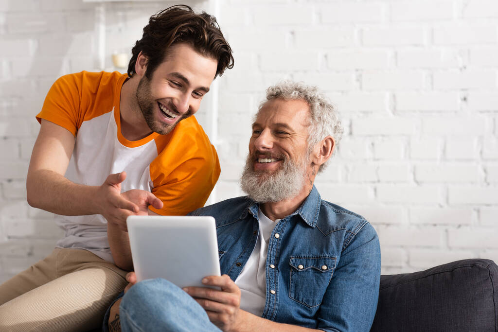 Sonriente hombre apuntando a la tableta digital cerca de padre maduro en casa  - Foto, imagen