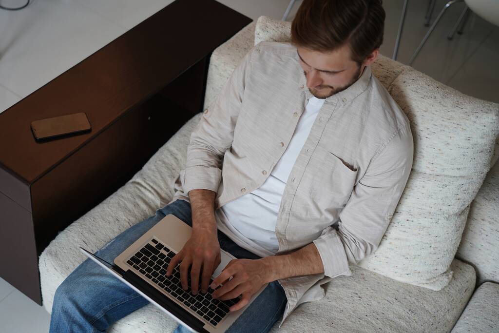 Uomo che utilizza il computer portatile, seduto sul divano a casa, chatta o fa shopping in internet, freelance che lavora online - Foto, immagini