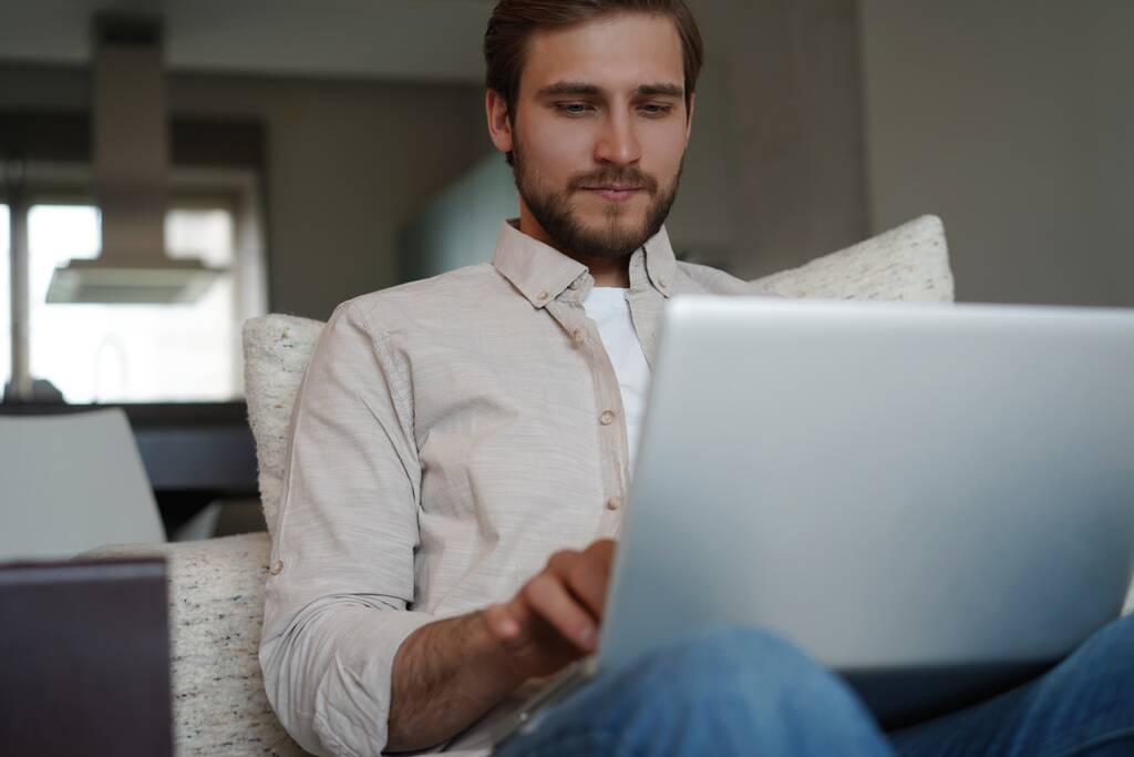 Mężczyzna korzystający z laptopa, siedzący na kanapie w domu, rozmawiający lub robiący zakupy w Internecie, wolny strzelec pracujący online - Zdjęcie, obraz