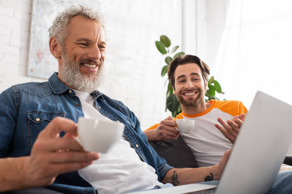 Усміхнений чоловік тримає каву біля сина і розмитий ноутбук
  - Фото, зображення