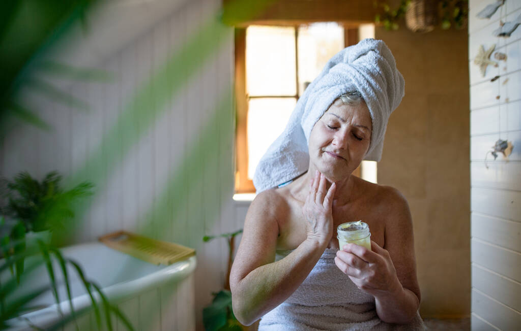 自宅のバスルームにタオルで包まれた幸せな先輩女性,自家製ボディ保湿剤を適用します. - 写真・画像
