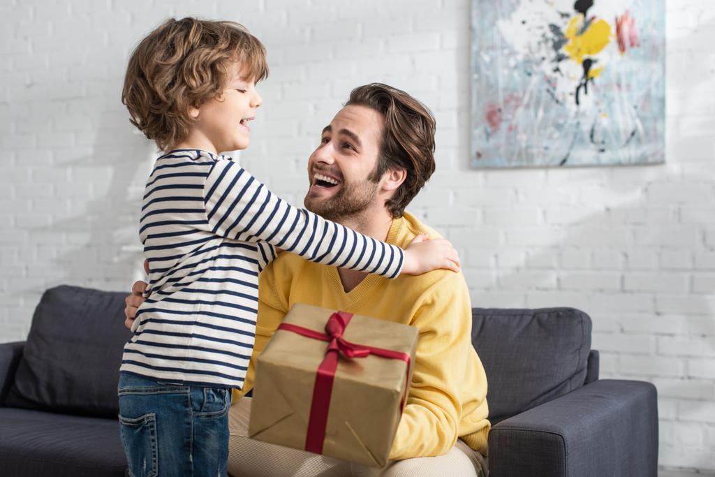 Cheerful kid hugging father with present on blurred foreground  - Photo, Image