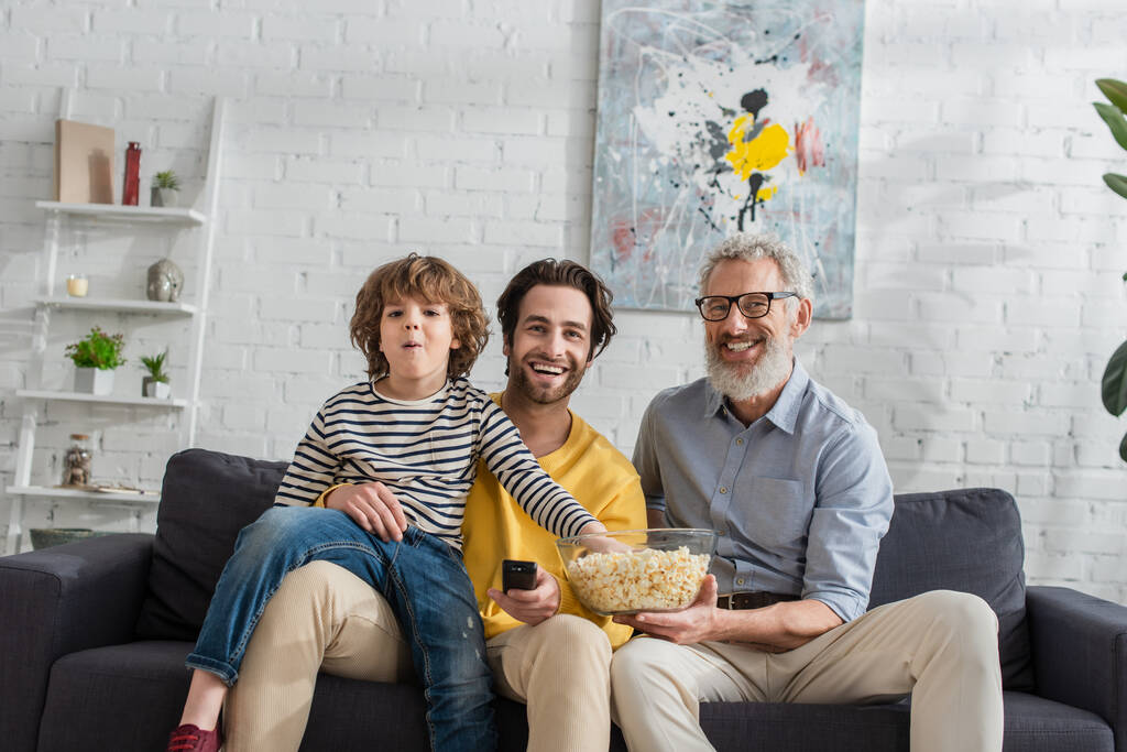 Sorridente nonno che tiene ciotola con popcorn vicino al figlio con telecomando e nipote  - Foto, immagini