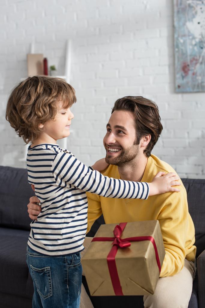 Χαμογελώντας πατέρας με κουτί δώρου κάθεται κοντά στο γιο στο σπίτι  - Φωτογραφία, εικόνα