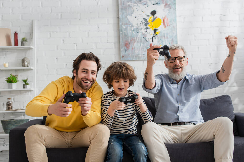 KYIV, UKRAINE - APRIL 12, 2021: Mature man showing yes gesture near son and grandson with joysticks  - Photo, Image