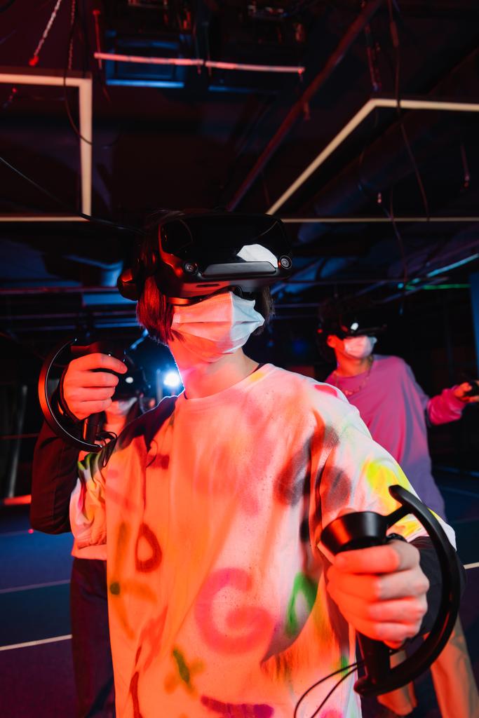 adolescent garçon dans le jeu de masque médical près des amis flous dans la zone de jeu vr - Photo, image