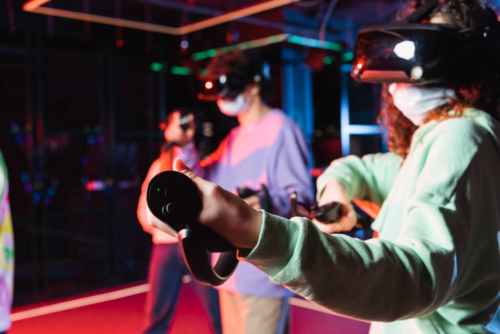 african american teenage girl in vr headset and safety mask gaming near blurred friends - Photo, Image