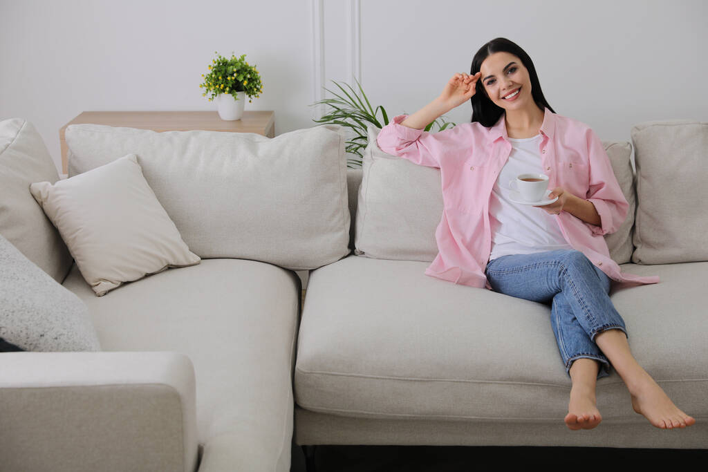 Женщина с чашкой напитка отдыхает на диване в гостиной - Фото, изображение