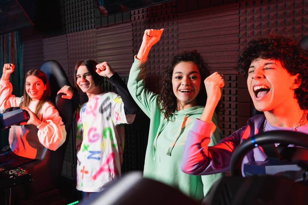 heureux adolescents multiethniques montrant geste de victoire près de simulateurs de voiture - Photo, image