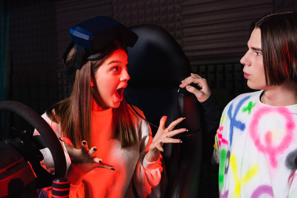 verbaasd meisje gebaren terwijl praten met vriend in de buurt auto simulator - Foto, afbeelding