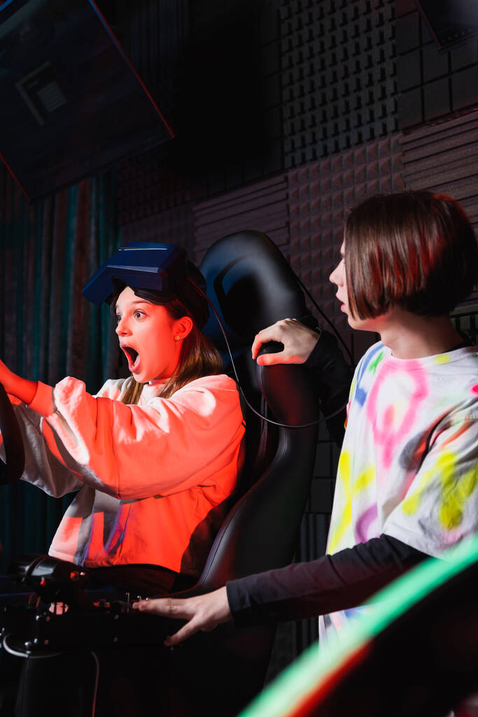 menina surpreso gritando enquanto correndo no simulador de carro perto de amigo adolescente - Foto, Imagem