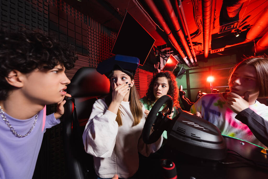 shocked girl on racing simulator covering mouth with hand near interracial friends - Photo, Image
