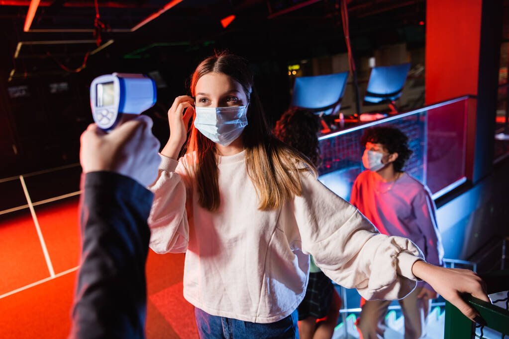 tizenéves lány orvosi maszk közelében vezérlő pirométer és barátai elmosódott háttér - Fotó, kép