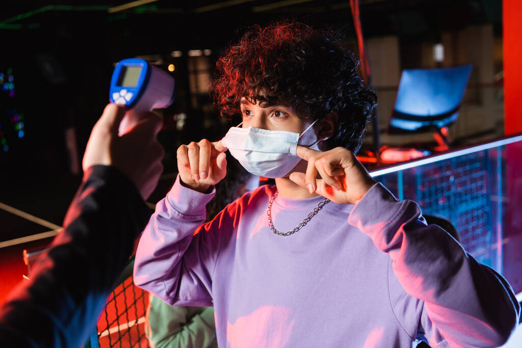 gamer adolescente ajustando máscara médica perto do controlador com pirômetro - Foto, Imagem