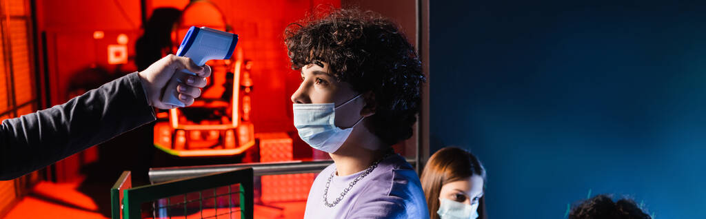 мальчик-подросток в медицинской маске рядом с контроллером с пирометром и девушкой на размытом фоне, баннер - Фото, изображение