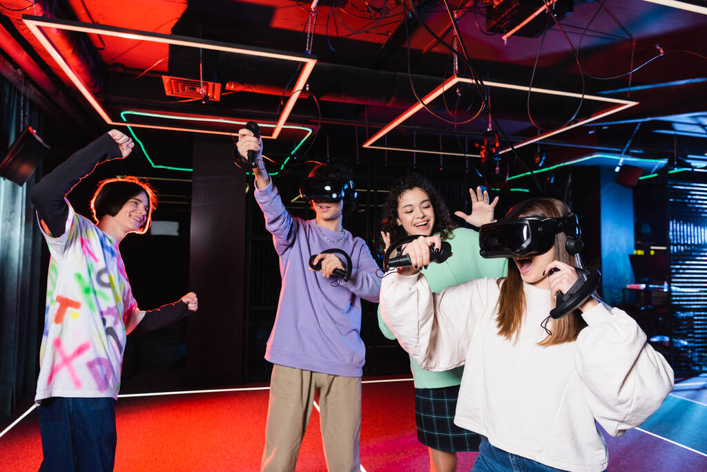 cheerful teenage boy showing win gesture near multicultural friends in vr play room - Photo, Image