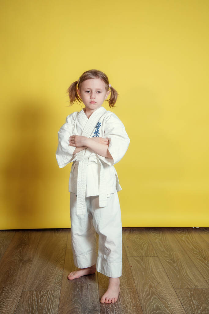 Porträt eines 5-jährigen kaukasischen Mädchens im Kimono, das zu Hause vor gelbem Hintergrund Karate übt - Foto, Bild
