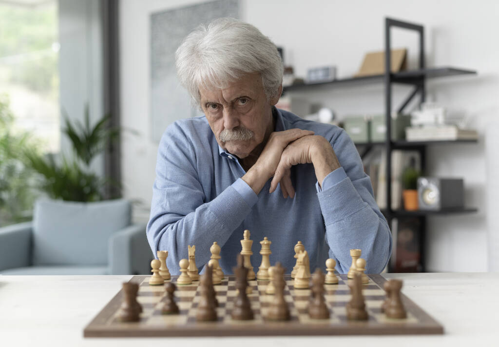 Впевнений старший чоловік грає в шахи вдома, він дивиться на камеру
 - Фото, зображення