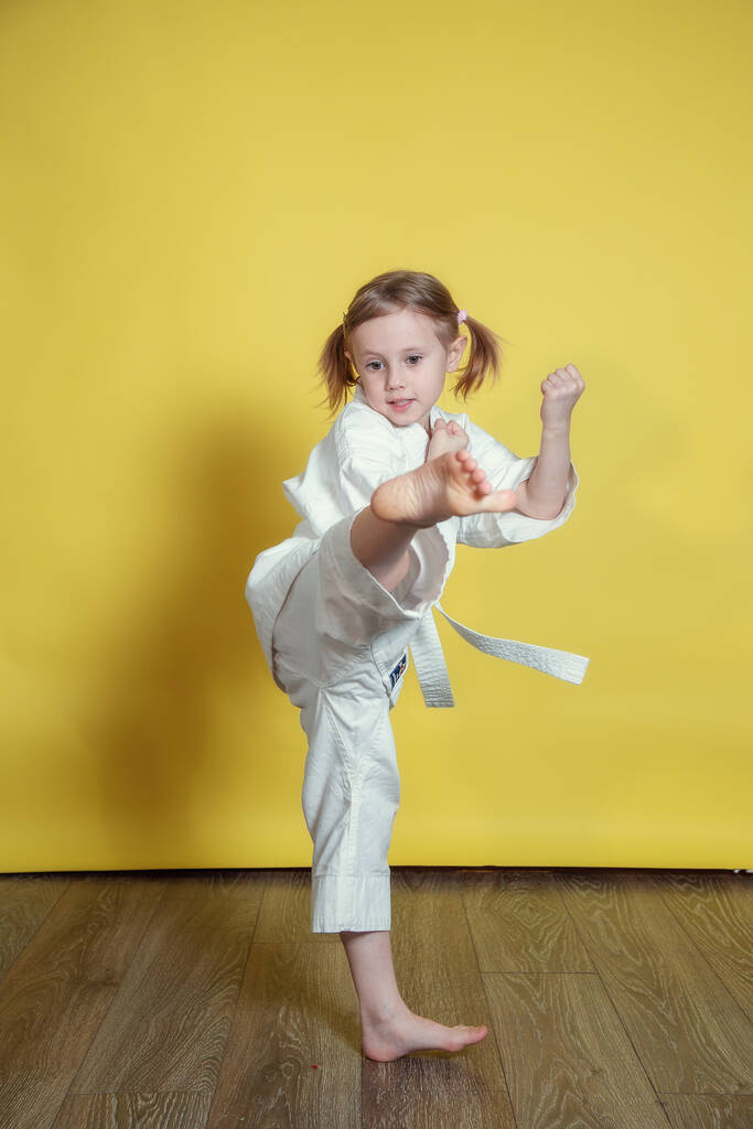 Портрет 5-летней белой девушки в кимоно, практикующей карате на жёлтом фоне дома - Фото, изображение
