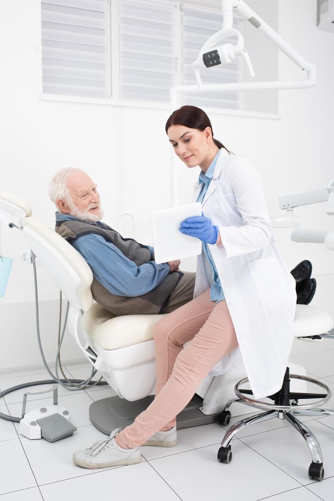 paciente mayor acostado en la silla dental y tomando la consulta del médico con el comprimido en las manos - Foto, Imagen