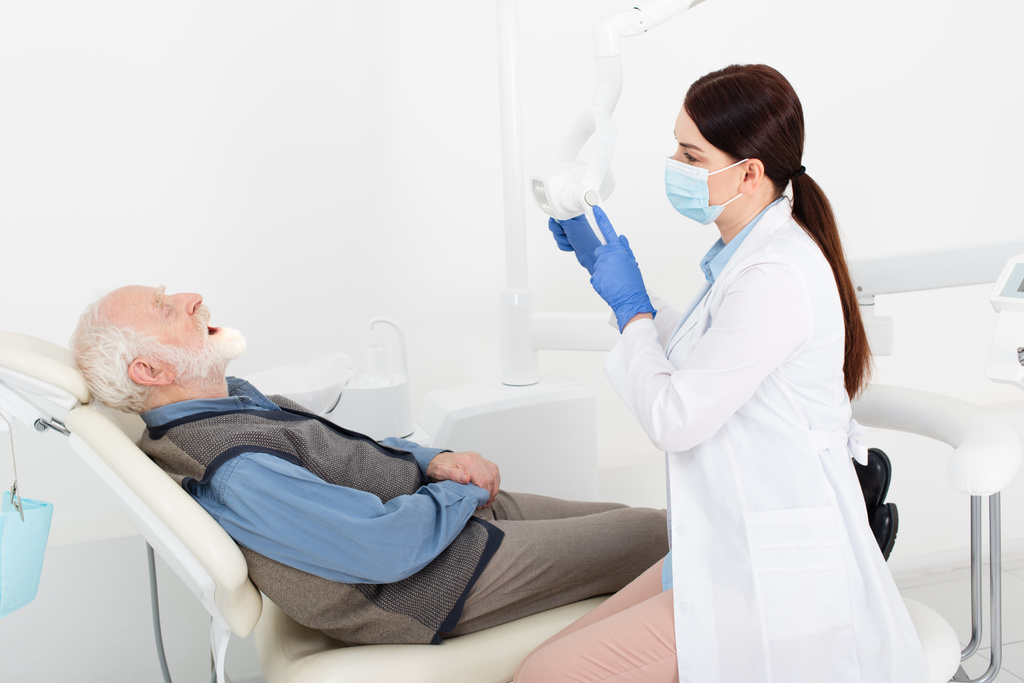 fogorvos orvosi maszkban és kék latex kesztyűben, orvosi lámpát érintve a fogorvosi székben ülő idős beteg előtt. - Fotó, kép