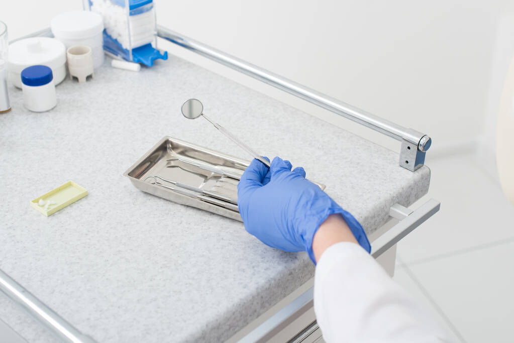 обрізаний вид рук стоматолога в латексній рукавичці, що бере дзеркало з металевого лотка на стіл у стоматологічній клініці
 - Фото, зображення