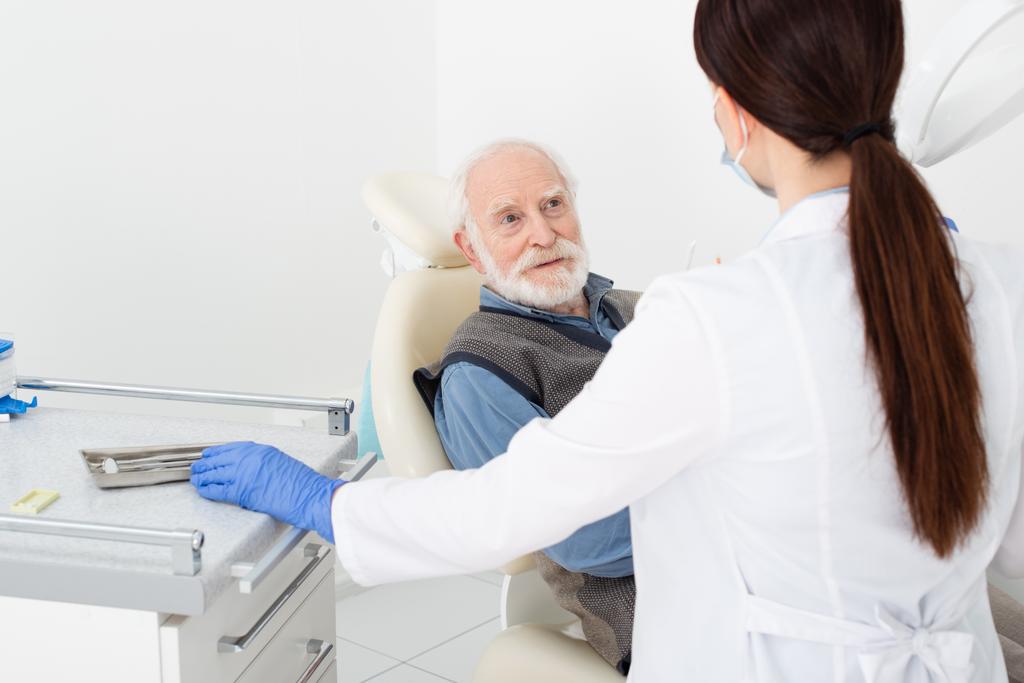 улыбающийся мужчина получает консультацию у стоматолога в латексных перчатках в стоматологической клинике - Фото, изображение
