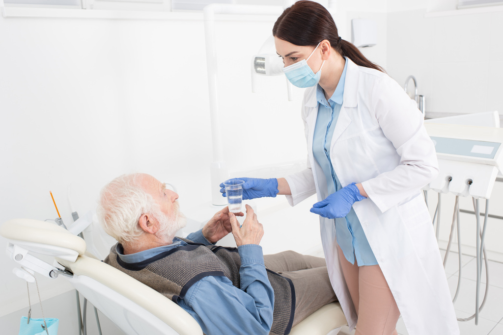 fogorvos ad egy pohár vizet egy idős betegnek, aki fogorvosi székben fekszik.  - Fotó, kép