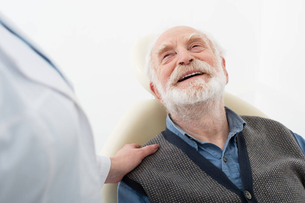 paciente anciano sonriente sentado en silla dental con dentista mano sobre hombro - Foto, Imagen